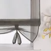 A close-up view of a gray sheer Roman shade with tie-up details, hanging in front of a window. Produktbild-Vorschau