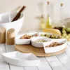 A white ceramic serving platter with multiple compartments filled with nuts and olives, placed on a wooden board on a tiled surface. In the background, there are salt and pepper shakers, a bowl with chopsticks, and bottles of oil. Náhled obrázku produktu