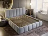 A modern bedroom with a light gray upholstered bed frame and a wooden slatted base. The bed is placed on a brown rug, with a large decorative moon artwork on the wall behind it. A bedside table with a vase and books is visible on the left, and a window wit Produktbild-Vorschau