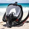 A black full-face snorkel mask is placed on a sandy beach with the ocean in the background. Podgląd zdjęcia produktu