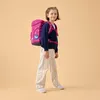 A young girl wearing a pink backpack and striped pants stands against a plain background. Produktbild-Vorschau