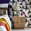 A bedside table with two drawers, a lamp, a vase with flowers, and decorative items, placed next to a bed with pillows and a blanket. The background features a patterned tile wall. Produktbild-Vorschau