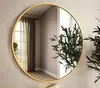 A round mirror with a gold frame hangs on a wall, reflecting a room with curtains and a plant.