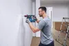 A man is drilling a hole in a white wall with a blue and black rotary hammer drill. The room appears to be under renovation, with plastic sheeting on the floor and a ladder in the background. Produktbild-Vorschau
