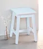 A white wooden stool is placed against a light-colored wall, with a vase of white flowers to its left. Produktbild-Vorschau