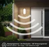 An outdoor wall lamp with motion sensor features is shown mounted on a wooden-paneled wall next to a door. The sensor's detection range is illustrated with curved lines. Anteprima dell’immagine prodotto
