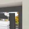 Close-up view of a white blind mechanism attached to a dark gray window frame, with a blurred background of trees and a street. Produktbild-Vorschau