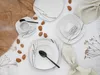 A flat lay of a white porcelain dinnerware set with a modern abstract design, arranged with almonds and a linen napkin on a white surface, with a textured brown paper element in the background. Náhľad obrázku produktu