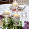 A table setting with floating candles in glass cylinders decorated with ribbons and pearls, surrounded by greenery and a purple cloth. Produktbild-Vorschau