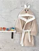 A child's bathrobe with a bunny hood hangs on a concrete wall next to a shelf with toys. Produktbild-Vorschau