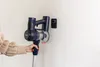 A hand is holding a cordless vacuum cleaner near a wall-mounted charging dock. Náhľad obrázku produktu