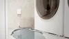 A white smart water sensor is mounted on a wall next to a washing machine, with water leaking onto the floor. Podgląd zdjęcia produktu