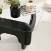 A close-up view of a modern coffee table with a glass top, showcasing a plant in a black vase and a lit candle on a book. Produktbild-Vorschau