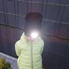 A child wearing a black beanie with a built-in light and a yellow jacket stands in front of a fence. Produktbild-Vorschau