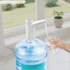 A white electric water dispenser pump is dispensing water from a large blue water jug into a clear glass held by a person. Podgląd zdjęcia produktu