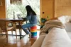 A woman is working on a laptop while sitting on a stack of colorful cushions in a wooden room with a large window overlooking greenery. Voir les images du produit
