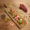 A wooden serving board with prosciutto, cherry tomatoes, basil, grapes, almonds, hazelnuts, and figs arranged on a wooden table. Anteprima dell’immagine prodotto
