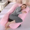 A woman is lying on a bed, using a large U-shaped pink pregnancy pillow. Produktbild-Vorschau