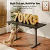 A cat is reaching up towards a standing desk with large gold letters spelling "70KG" on top. The desk is in a room with blinds and a plant. Produktbild-Vorschau