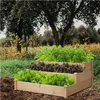 A tiered wooden raised garden bed filled with various types of lettuce and herbs, set outdoors in a garden with trees and foliage in the background. Produktbild-Vorschau