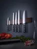 A set of knives mounted on a magnetic strip on a dark wall, with some food items in the foreground. Produktbild-Vorschau