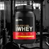 A tub of Optimum Nutrition 100% Whey Gold Standard protein powder is displayed in a gym setting. Produktbild-Vorschau