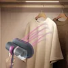 A hand holding a handheld vacuum cleaner, steaming a beige sweatshirt hanging in a closet. Produktbild-Vorschau