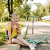 A young girl is sitting on a curb next to a scooter in a park. Podgląd zdjęcia produktu