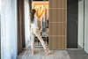 A woman is standing in front of an open closet, choosing clothes. The closet has a slatted wood design on the doors and is illuminated from within. Podgląd zdjęcia produktu