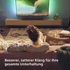 A person is relaxing on a couch watching TV, with a soundbar placed below the TV. Voir les images du produit