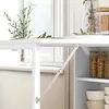 A close-up view of a white table with a metal support bracket. Shelves with food items are visible to the right. Náhľad obrázku produktu
