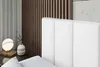 A close-up view of a white upholstered headboard of a bed, with a bedside table and lamp in the background. Produktbild-Vorschau