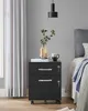 A black filing cabinet with two drawers is placed next to a bed with gray bedding. A vase with greenery and a mug are on top of the cabinet. Produktbild-Vorschau