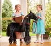 Two young girls in dresses standing with a black toy horse on wheels indoors, with a garden visible through a doorway. Náhľad obrázku produktu