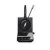 A black wireless headset with a microphone boom is placed on its charging base. The base has silver accents and several indicator lights. A small USB dongle is plugged into the side of the base. Podgląd zdjęcia produktu