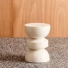A cream-colored, hourglass-shaped side table or stool is shown against a wooden panel background and a speckled floor. Produktbild-Vorschau