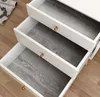 Three open drawers of a white dresser with a wood-grain liner. A rolled-up magazine and a rug are visible on the floor to the left. Náhľad obrázku produktu
