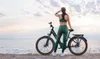 A woman in athletic wear stands next to an e-bike on a beach at sunset. Produktbild-Vorschau