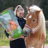 A young woman is holding a bag of horse feed next to a horse. Voir les images du produit