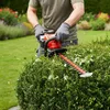 A person is using a red and black hedge trimmer to trim a green bush in a garden. Produktbild-Vorschau