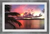 A framed picture of a sunset over the ocean with palm fronds in the foreground. Produktbild-Vorschau
