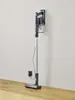 A cordless vacuum cleaner is mounted on a wall charging station next to a power outlet. The floor is made of wood. Voir les images du produit