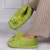 A person is holding up a Grinch slipper while wearing another one on their foot. Produktbild-Vorschau