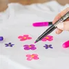 A hand is using a black fabric marker to draw a purple flower on a white t-shirt. Several other colorful flowers have already been drawn on the shirt. Produktbild-Vorschau