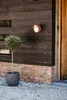A dark gray planter with a small green tree sits on gravel next to a brick wall and a dark wooden wall. An orange light fixture is mounted on the wooden wall. Voir les images du produit