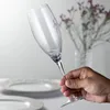 A hand holding a champagne flute with plates and flowers in the background. Produktbild-Vorschau