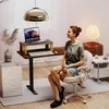 A woman is sitting at a standing desk in a home office setting. Produktbild-Vorschau