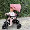 A baby sitting in a pink tricycle outdoors, with a hedge in the background. Produktbild-Vorschau