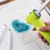 A hand is using a green airbrush tool to spray blue color onto a white stencil with a dinosaur shape. Produktbild-Vorschau