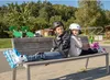 Two children wearing roller skates and helmets sit on a park bench. Produktbild-Vorschau